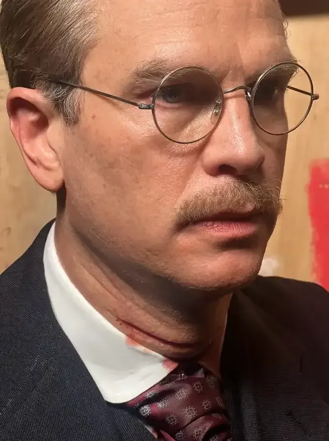 Man in glasses, mustache, and suit, with a cut throat, blood visible.
