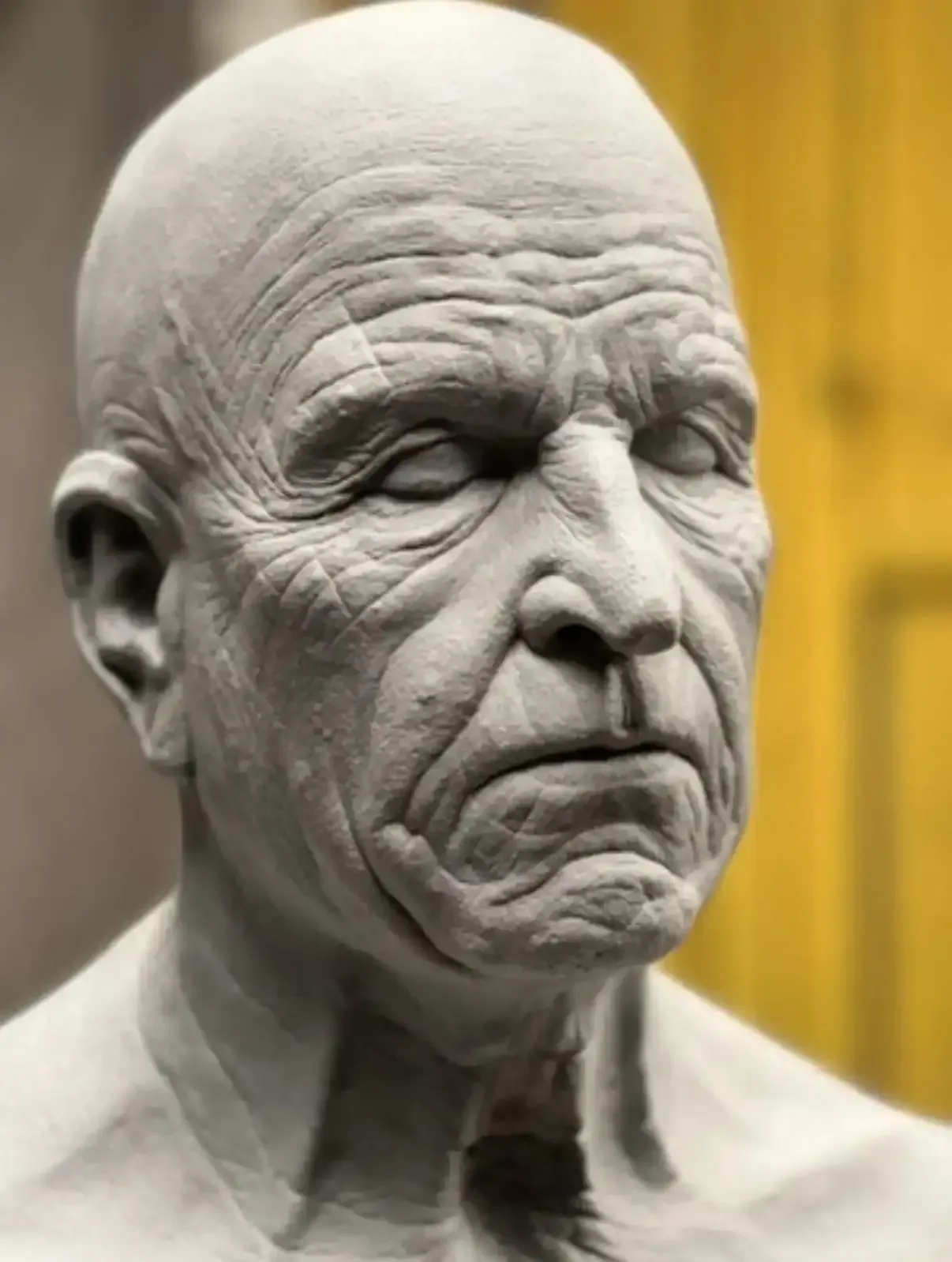 A textured, grey clay sculpture of a human head with prominent facial wrinkles and a serious expression.