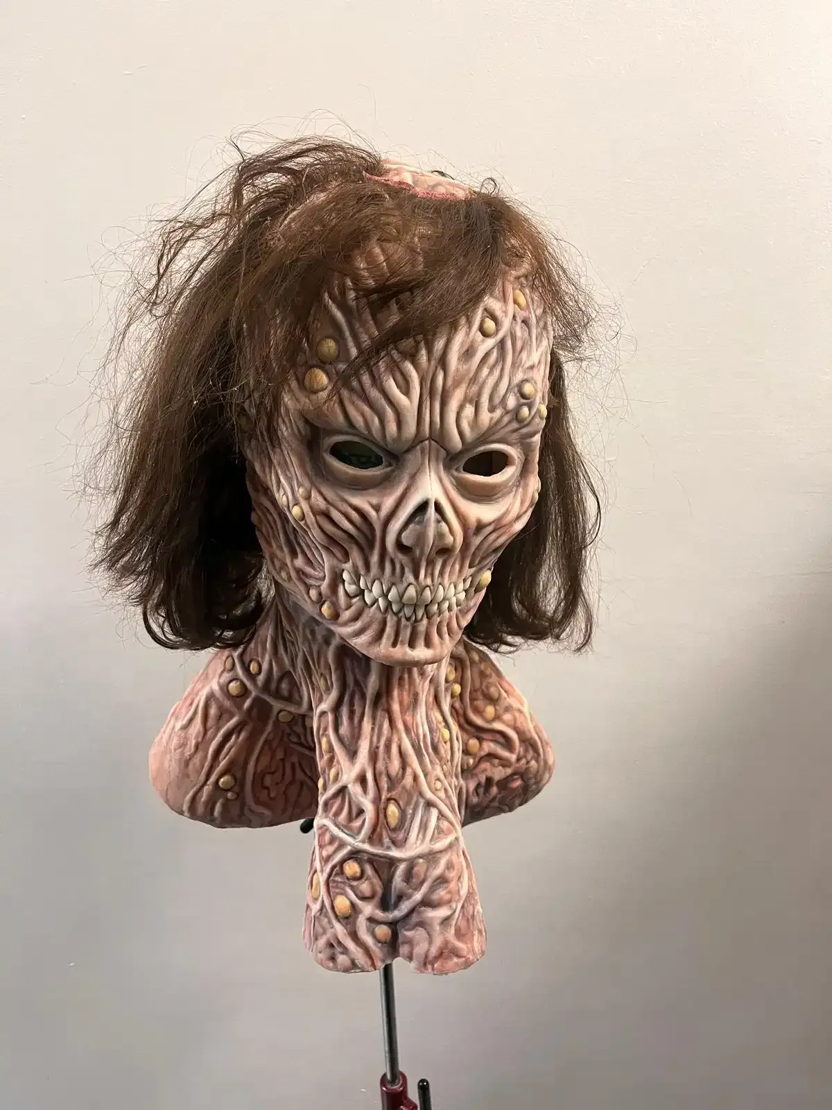 A grotesque, textured horror mask with brown hair, bulging skin patterns, and bared teeth mounted on a display stand.