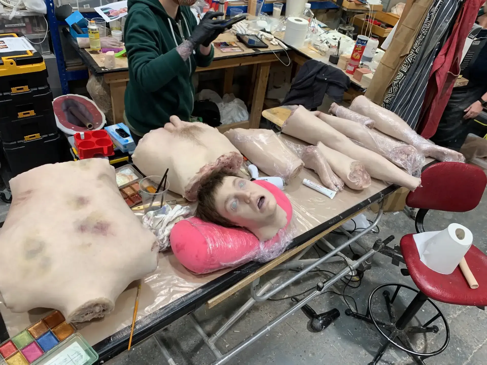 A table displays film prop body parts: torso, limbs, and a head. A person applies makeup, in a workshop.