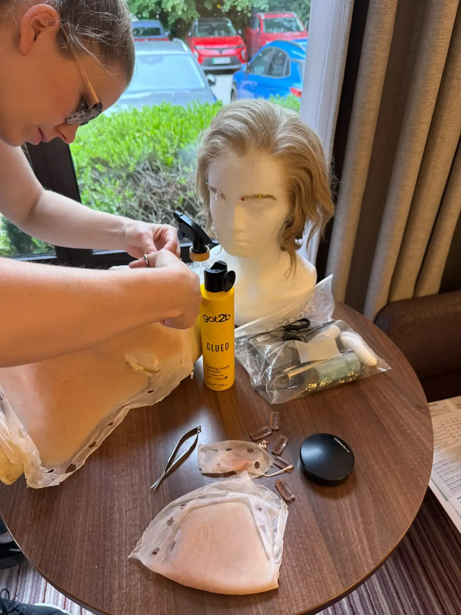 Person in white outfit has makeup applied by another with red apron, in workshop with masks.