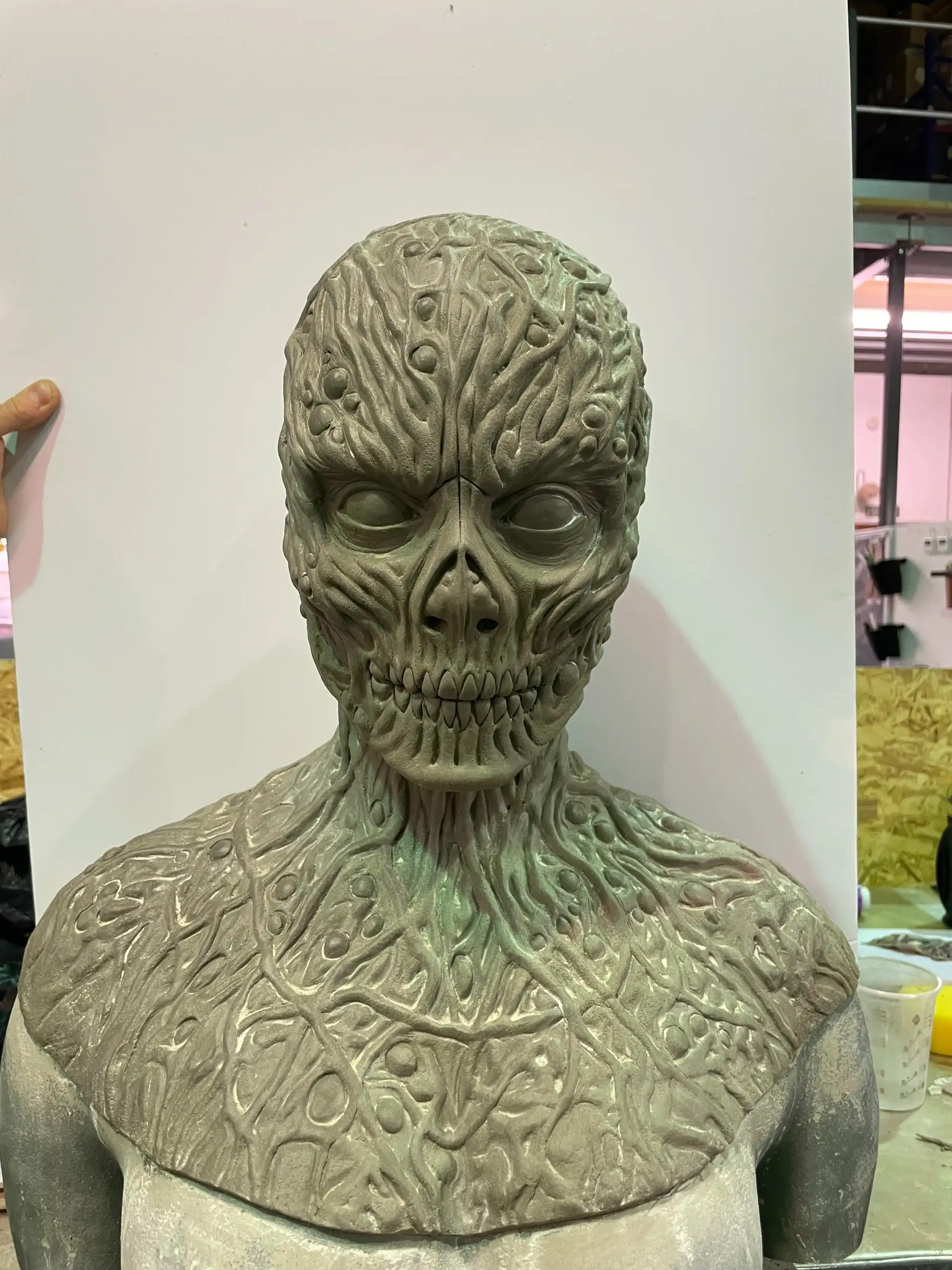 A detailed, green foam mask with pointed ears, textured facial features, and a beard, displayed on a workbench.