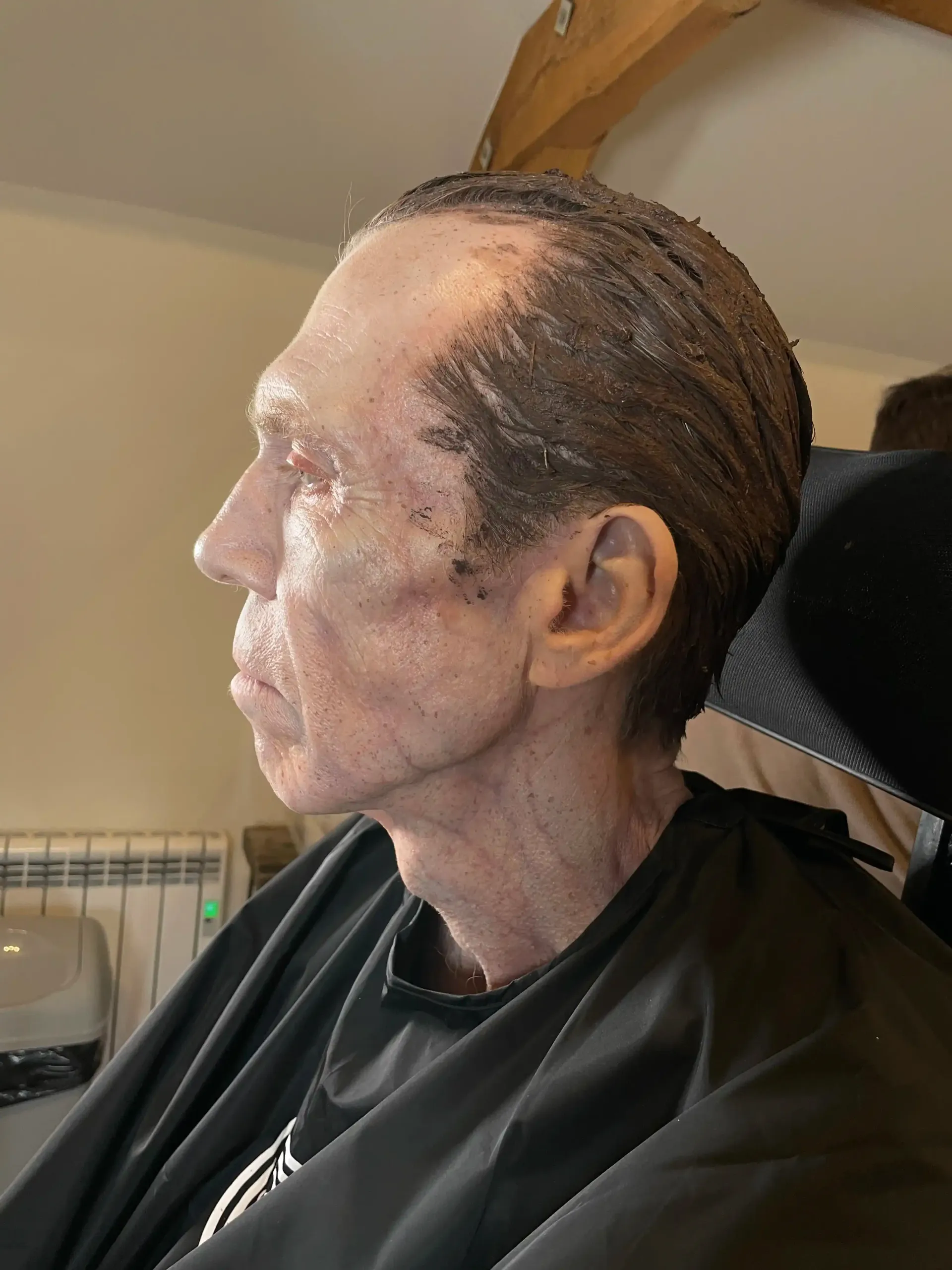 Man with skin makeup applied, in a salon setting. Side profile shows a weathered, textured appearance, with a black cape.