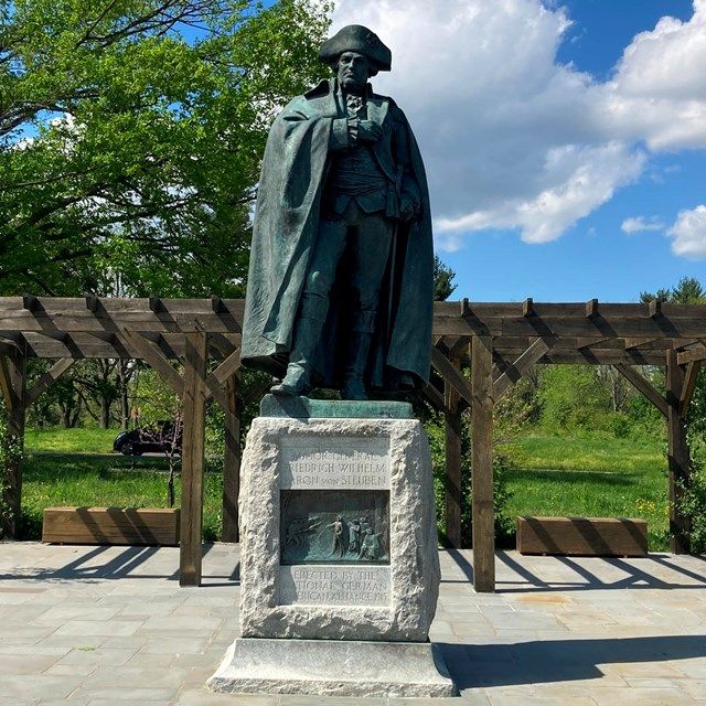 Bronze statue on a stone pedestal in a sunny park, with trees, wooden trellis, and blue sky behind it