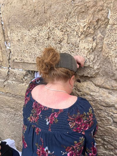 Person in a floral shirt and gray hat facing a stone wall, with head resting against it. Praying at Western Wall.