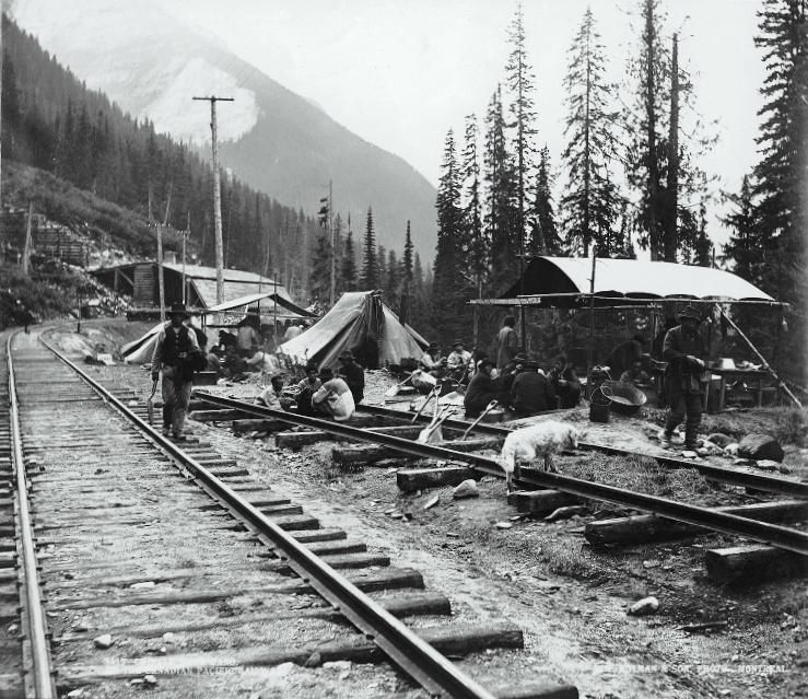 Mountain railroad camp beside tracks, with tents and workers in a black-and-white scene.