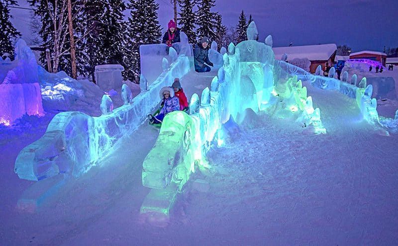 Illuminated ice sculpture display in a snowy outdoor park at dusk, glowing blue and purple