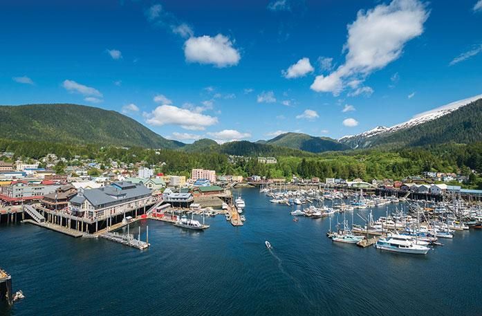 Marina with docked boats beside waterfront buildings, surrounded by mountains under a blue sky