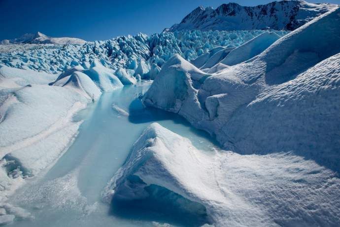 Blue glacier ice field with crevasses and snowy mountains in the background