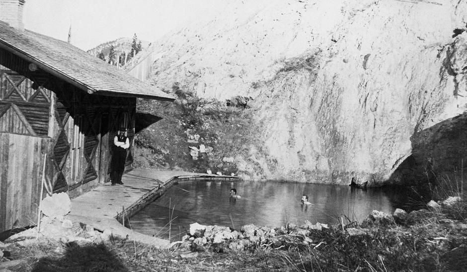 Black-and-white scene of a small wooden building beside a steaming hot spring pool in a rocky landscape