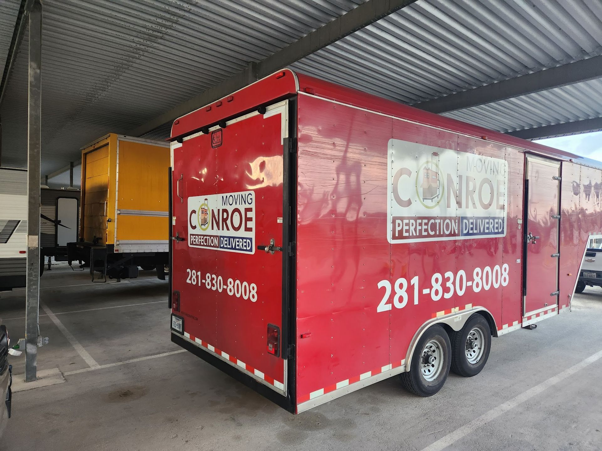 Red moving trailer parked under a covered area, with a yellow truck behind it.