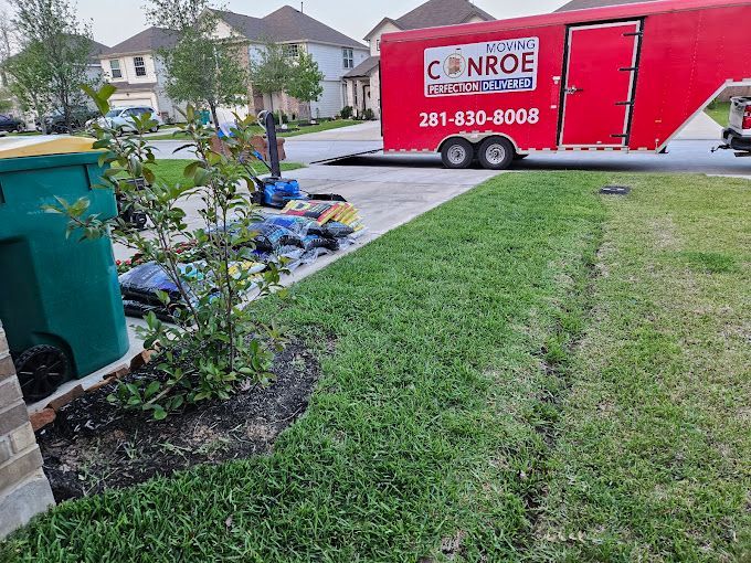 Red CanRoe trailer parked by a front yard with yard waste and a green trash bin nearby