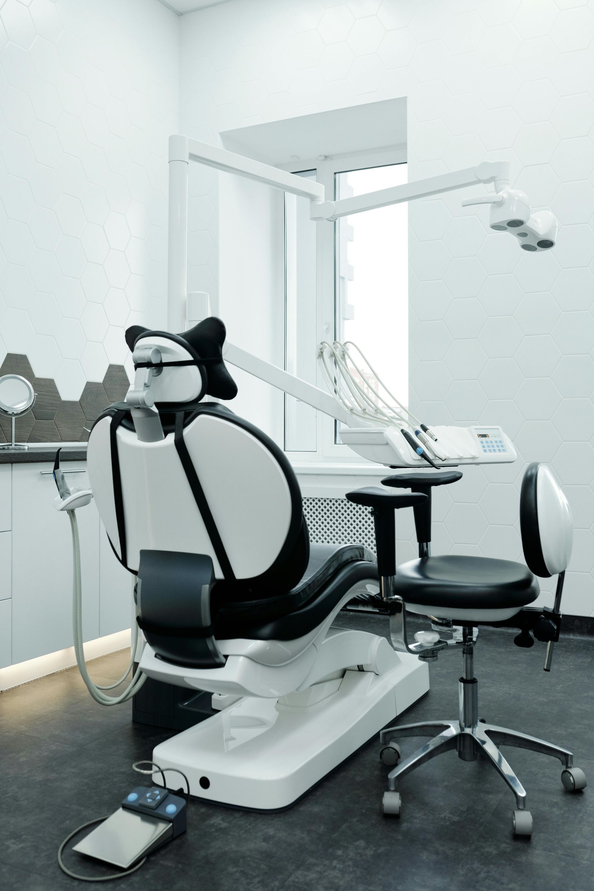 Dental exam chair in a bright clinic room with monitor, tray, and foot pedal