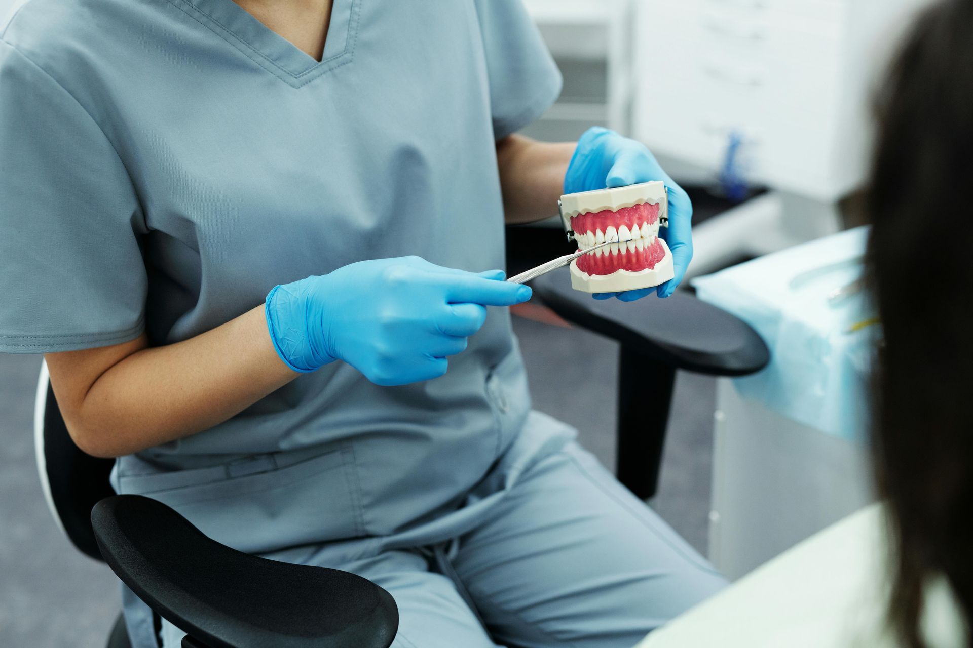 Dentist in gray scrubs holding a dental model and pointing with a blue gloved hand in a clinic