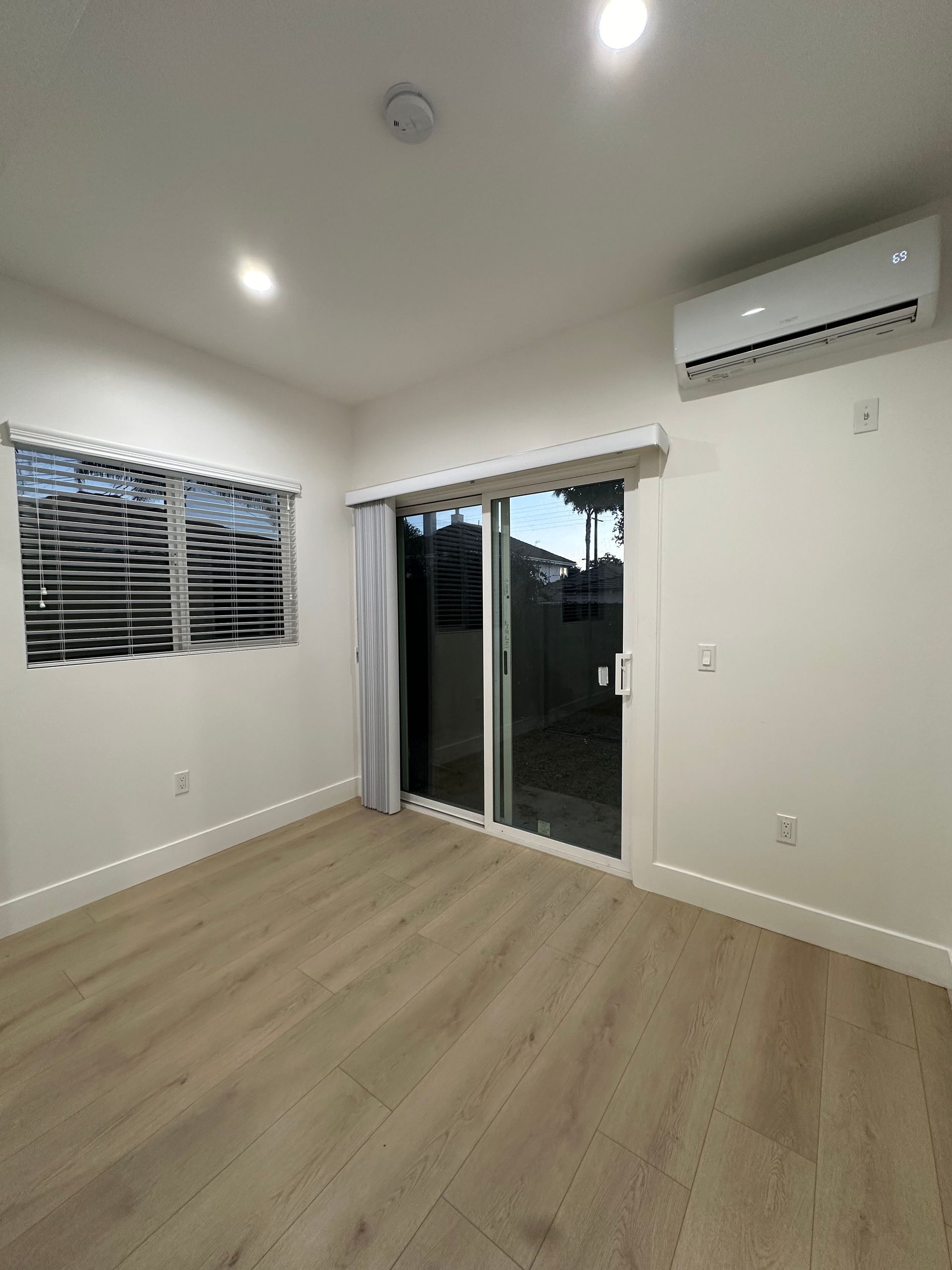 Empty modern room with light wood floor, white walls, window blinds, and sliding glass door.