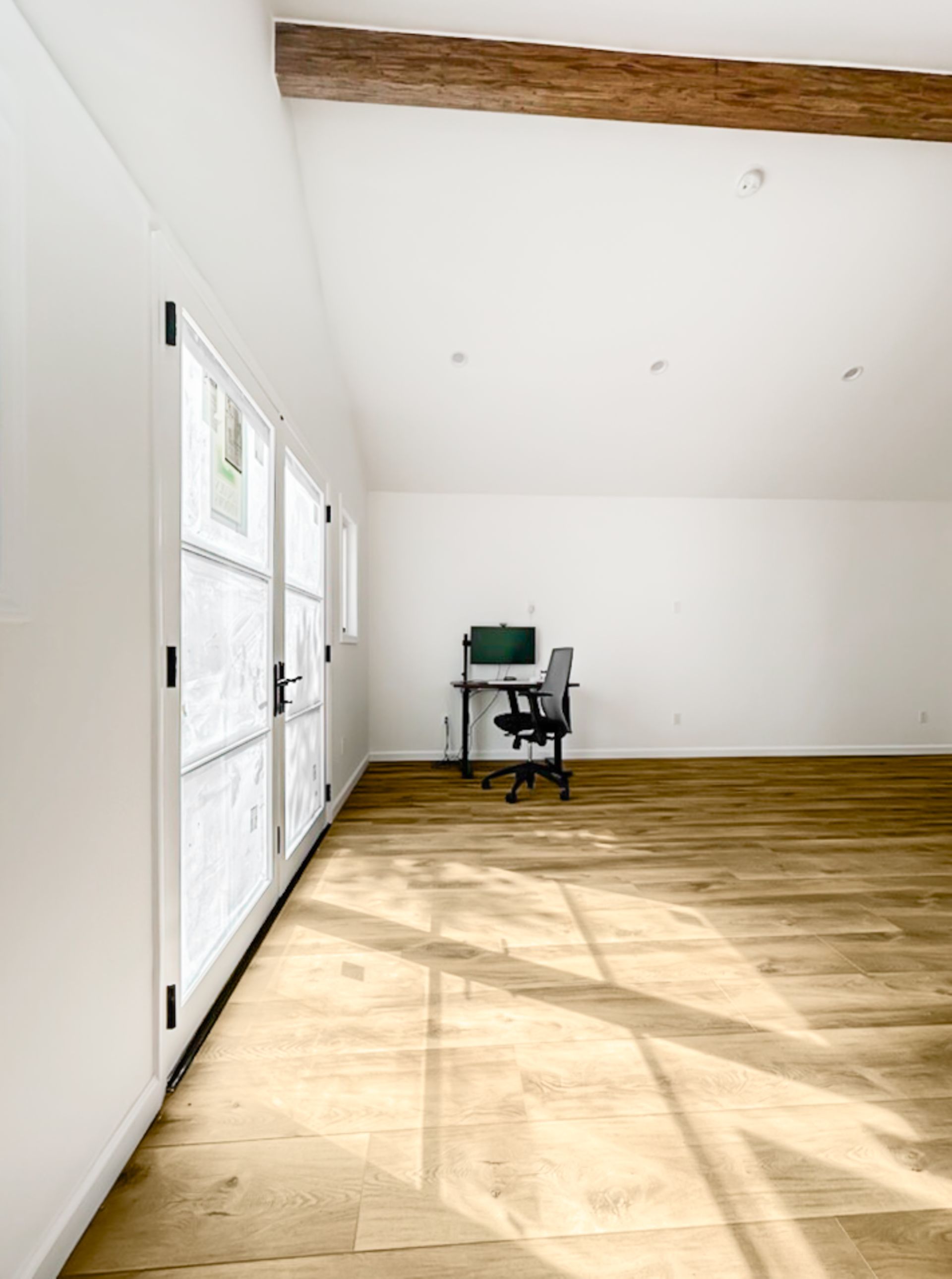Bright empty room with wood floor, white walls, large windows, and a desk with a computer near the back wall