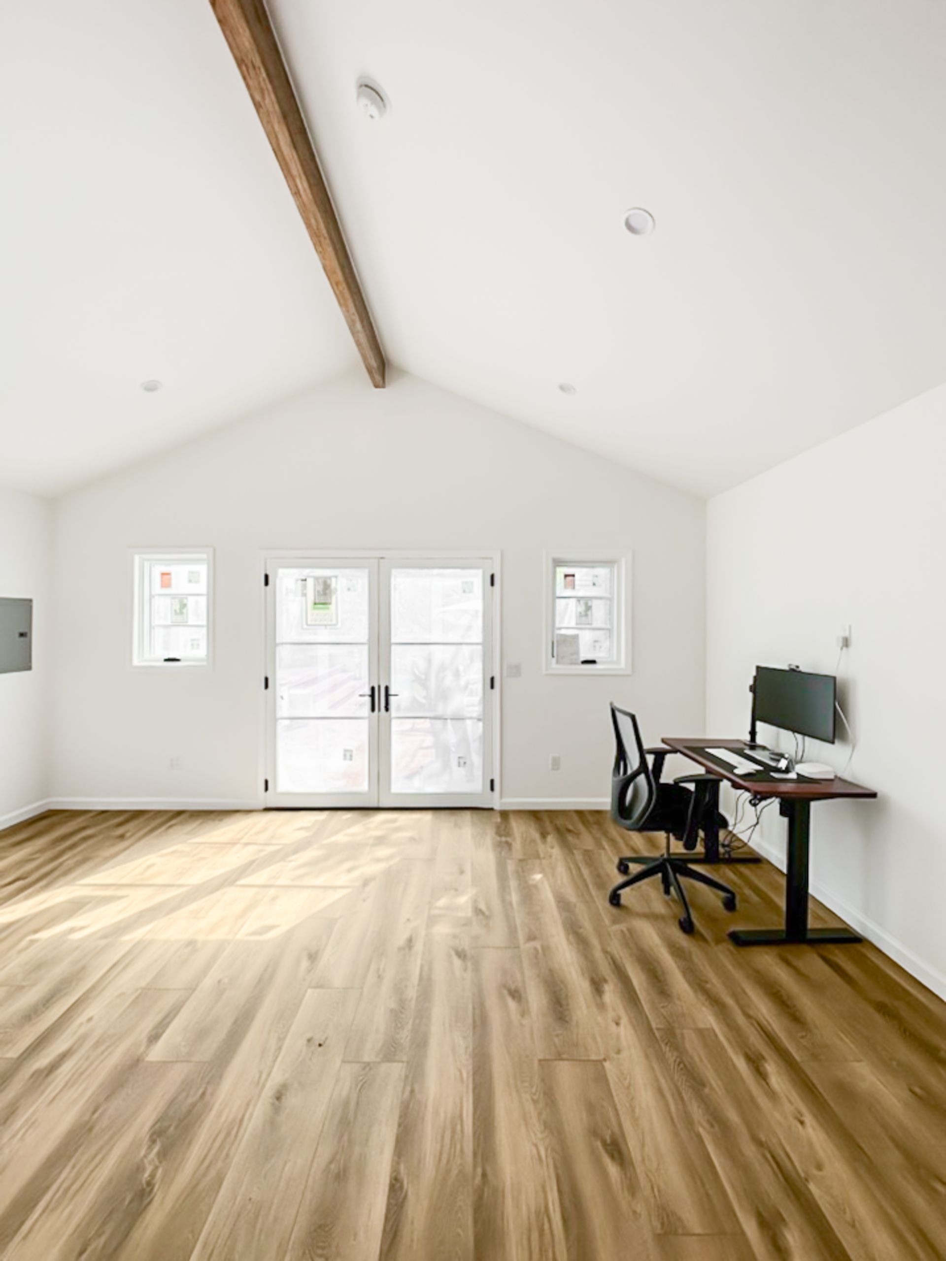 Bright empty room with wood floor, white walls, exposed beam, and a desk setup by the window.