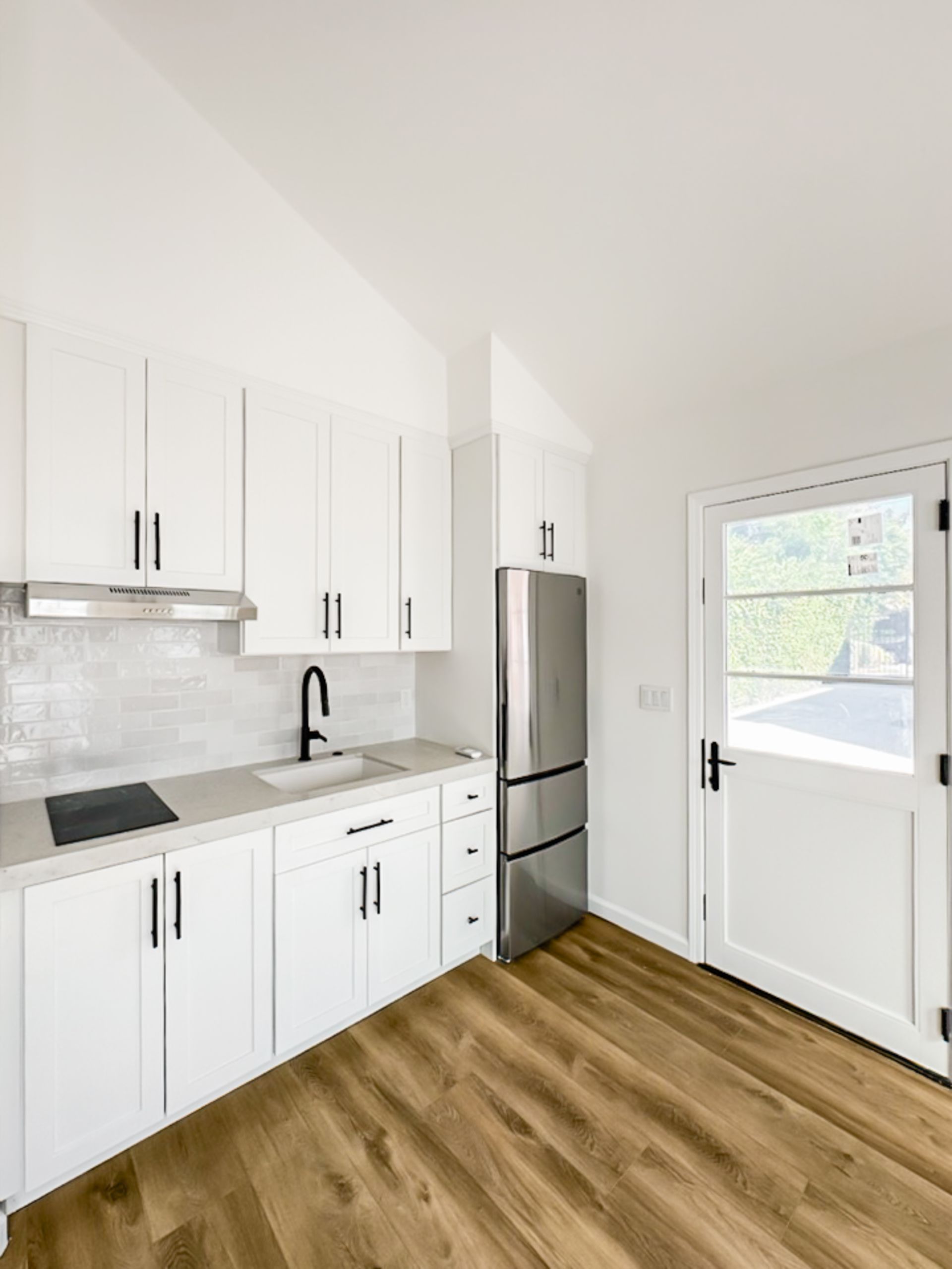 Bright kitchen with white cabinets, stainless fridge, black sink, and wood-look flooring beside a glass door