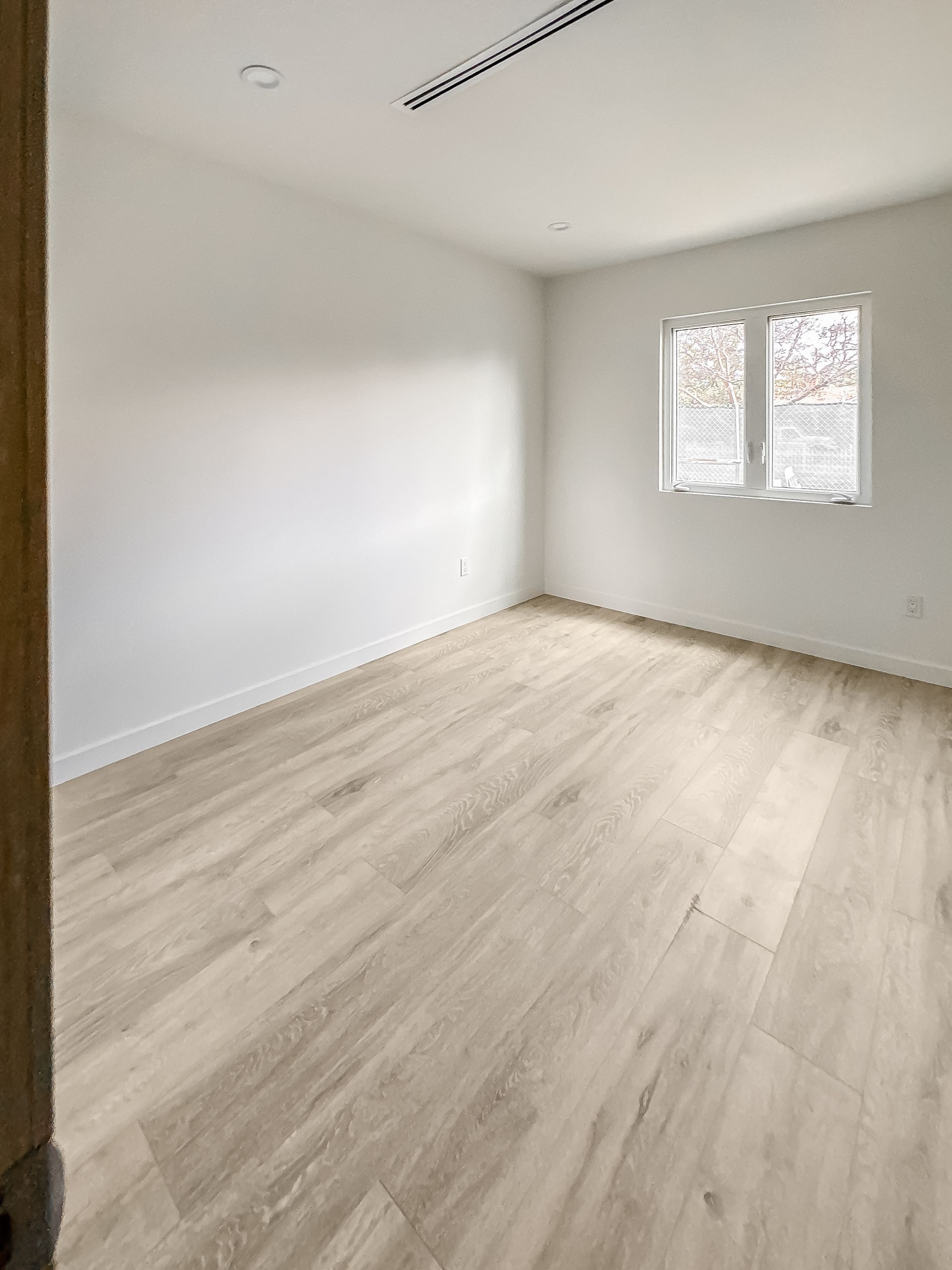 Empty light-filled room with white walls, pale wood floor, and a window on the far wall.