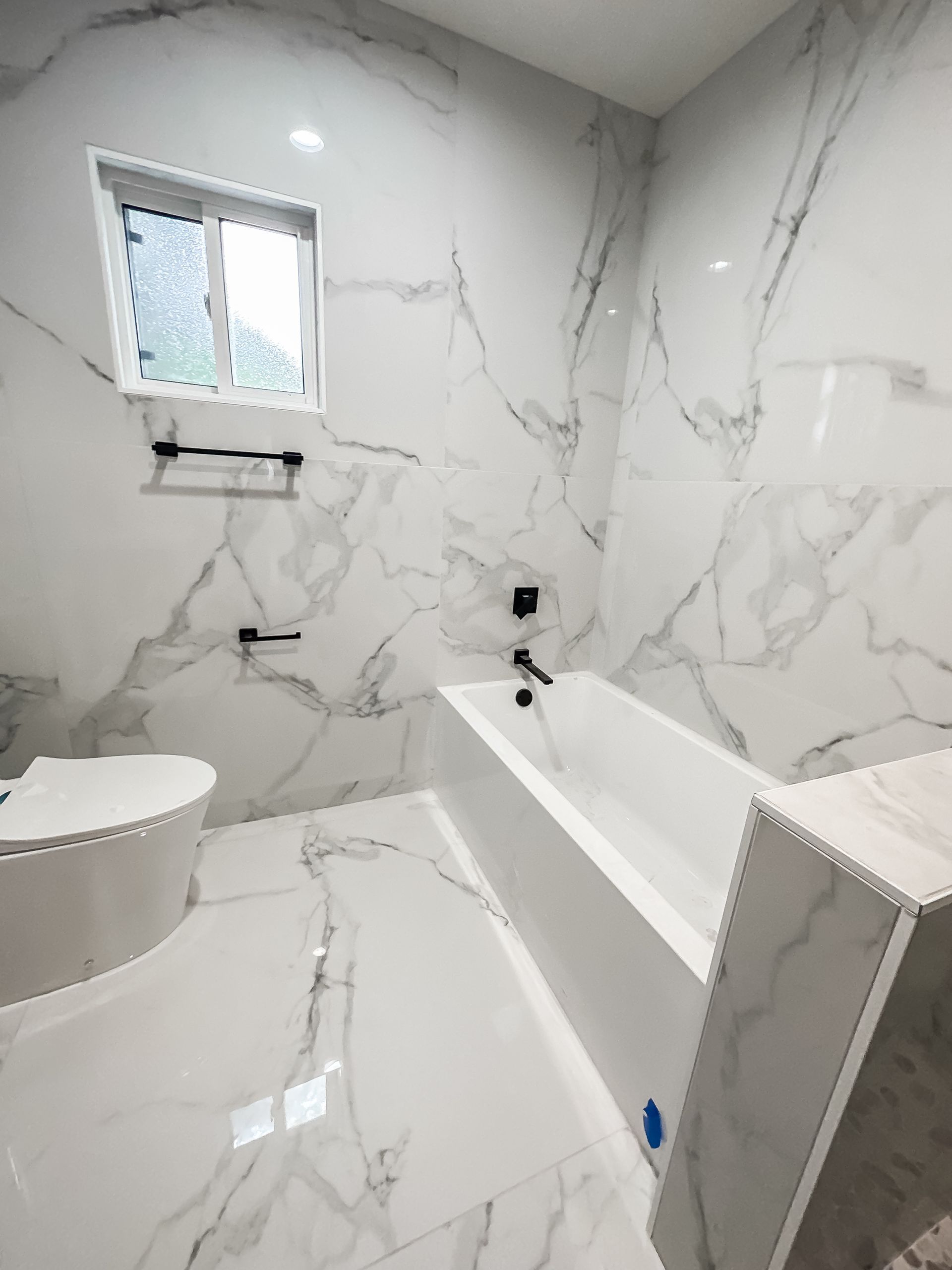 Modern white marble-tiled bathroom with a window, bathtub, and toilet.