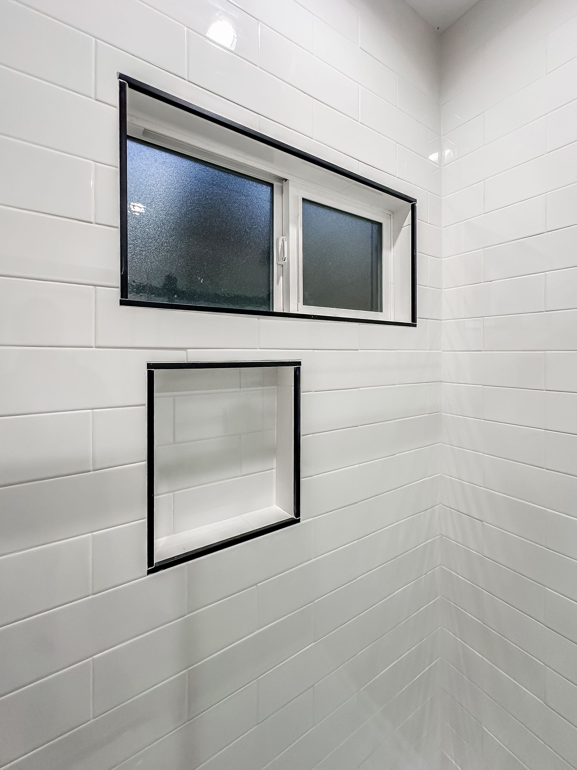 White tiled shower wall with two small frosted windows and a square recessed opening below