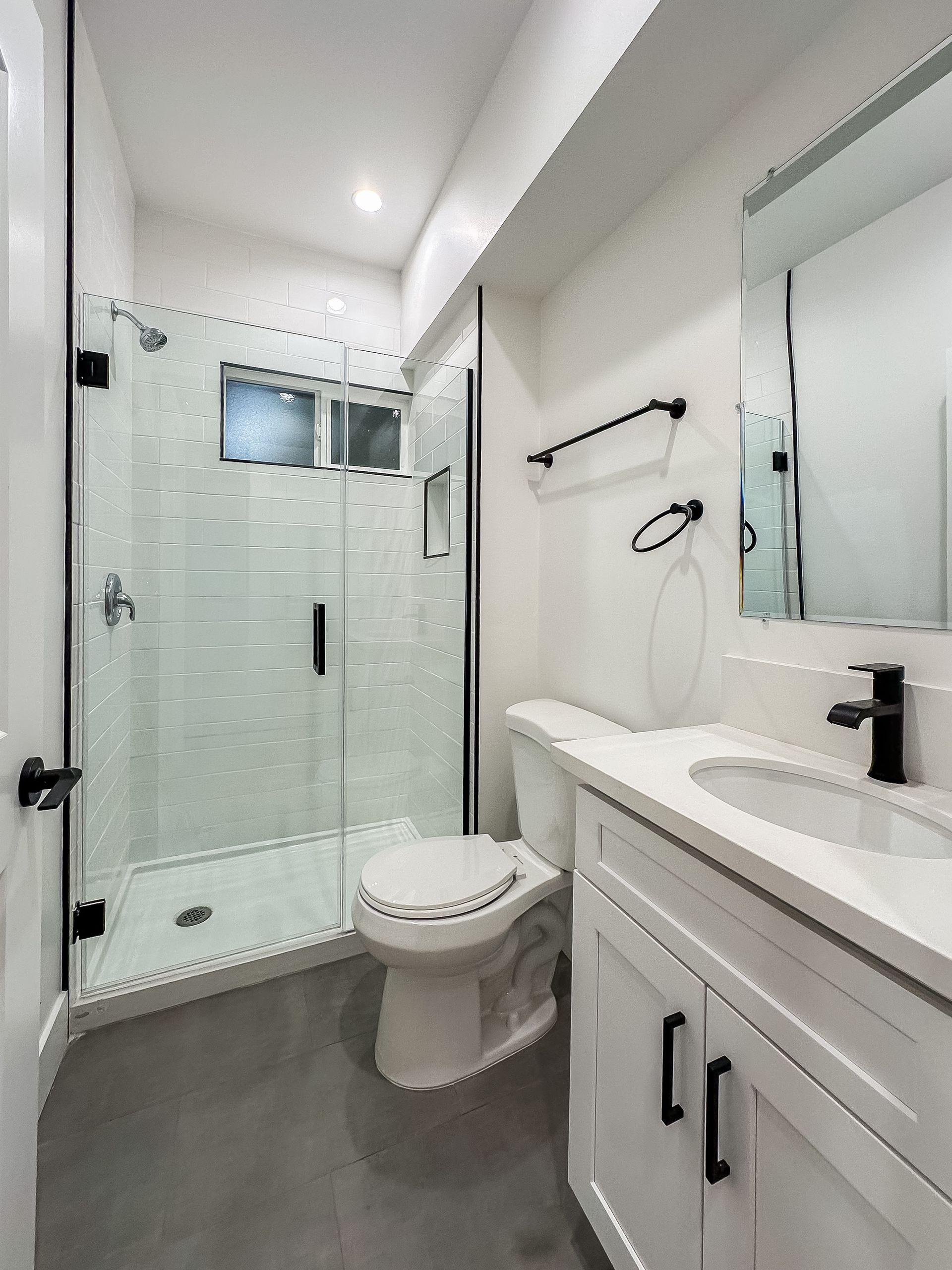 Narrow modern bathroom with white vanity, toilet, glass shower, and gray floor tiles