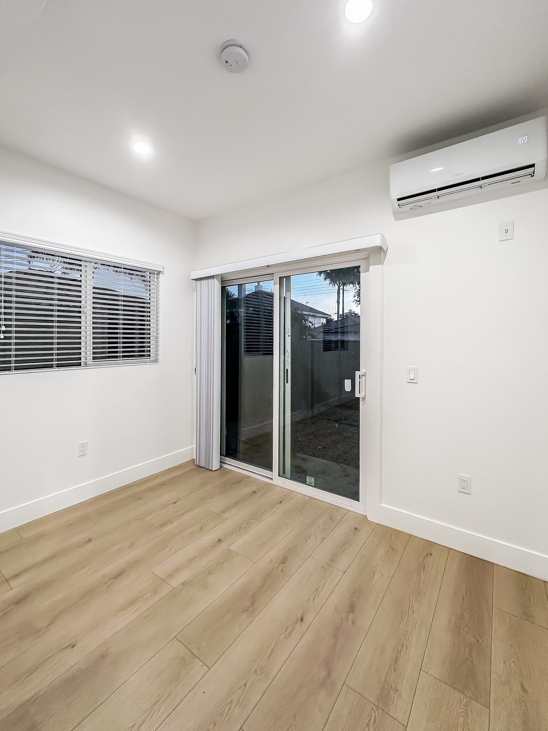 Empty bright room with wood floors, white walls, windows, sliding glass door, and wall-mounted air conditioner