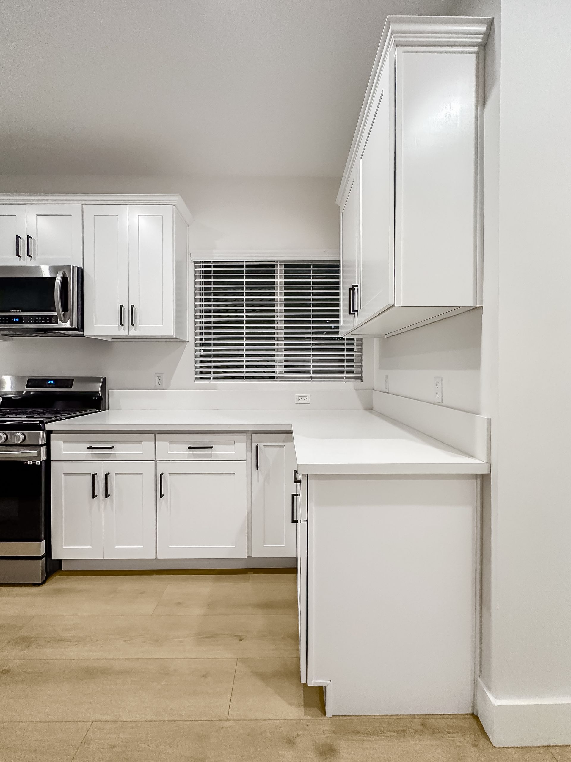 Bright white kitchen with cabinets, stove, microwave, window blinds, and tan floor