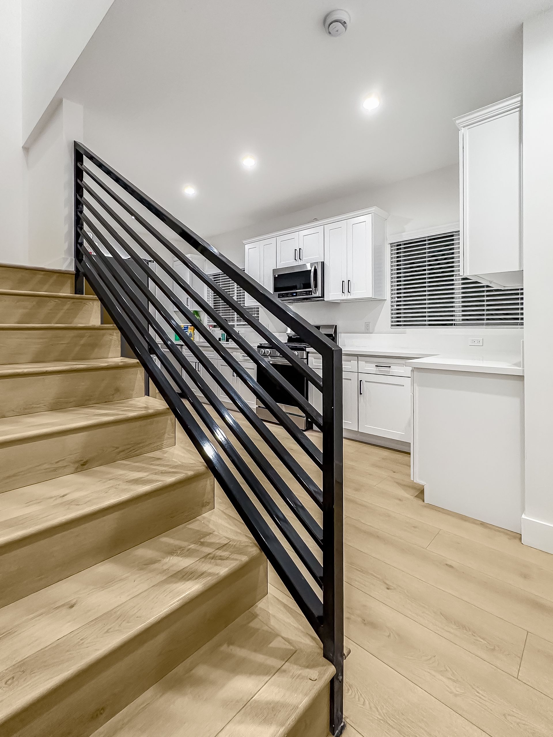 Modern kitchen with white cabinets, light wood floors, and a black staircase railing in the foreground