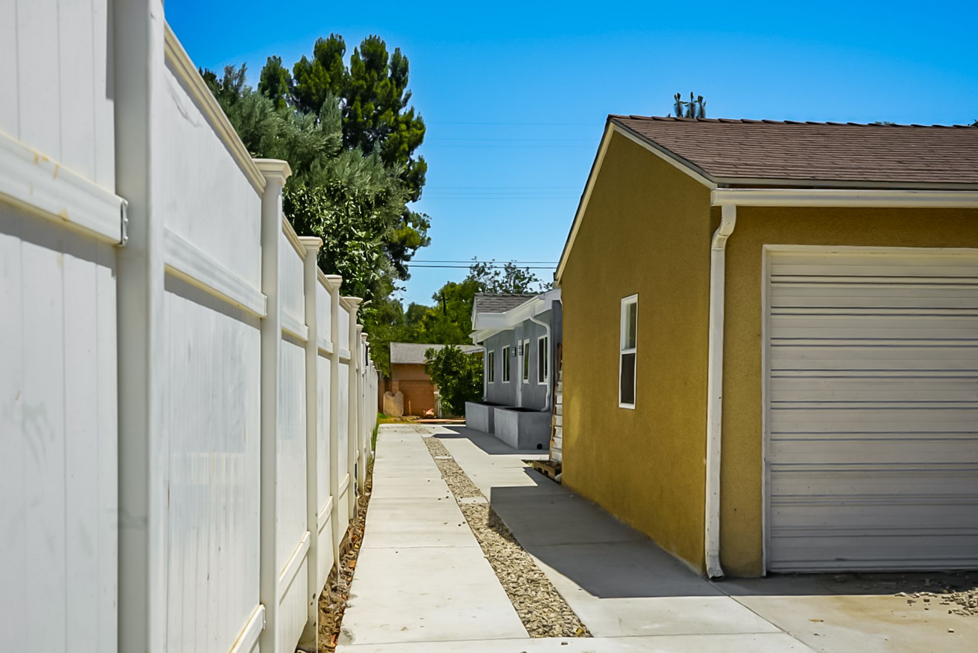 Narrow walkway between a white fence and yellow garage under a bright blue sky