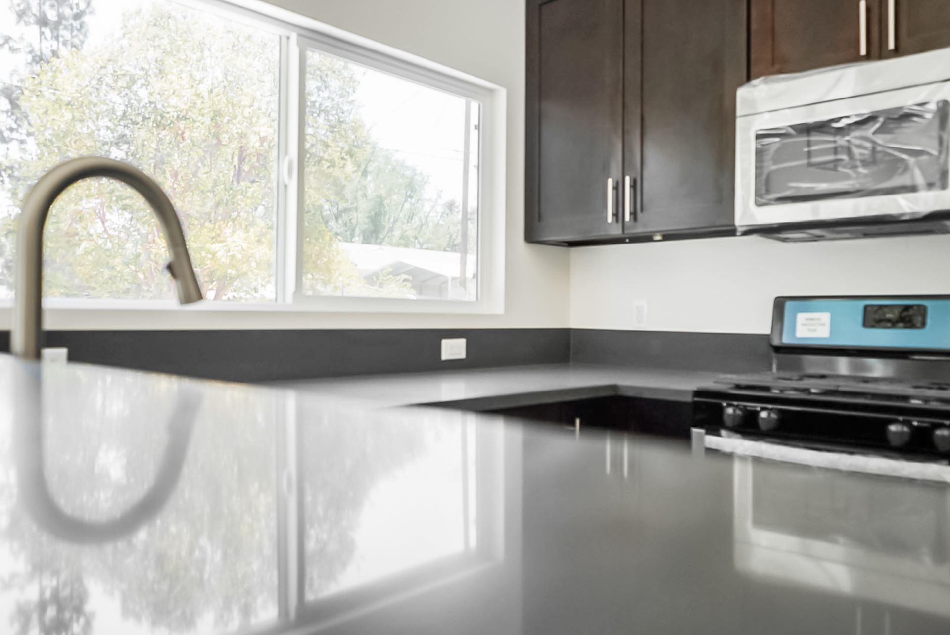 Modern kitchen with glossy black countertop, window, sink, dark cabinets, and stainless stove and microwave