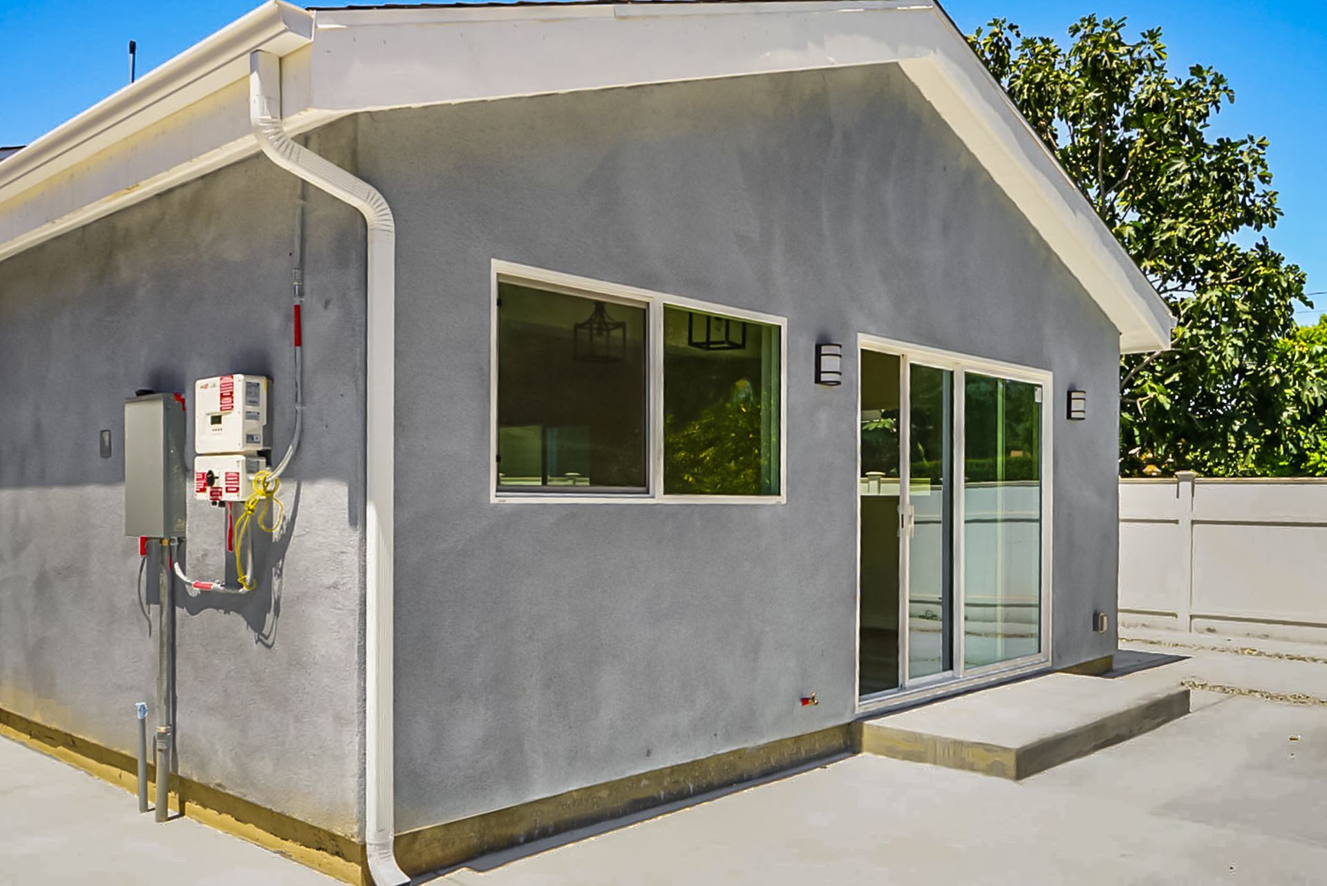 Small gray utility building with white trim, windows, and a glass door on a sunny paved lot.