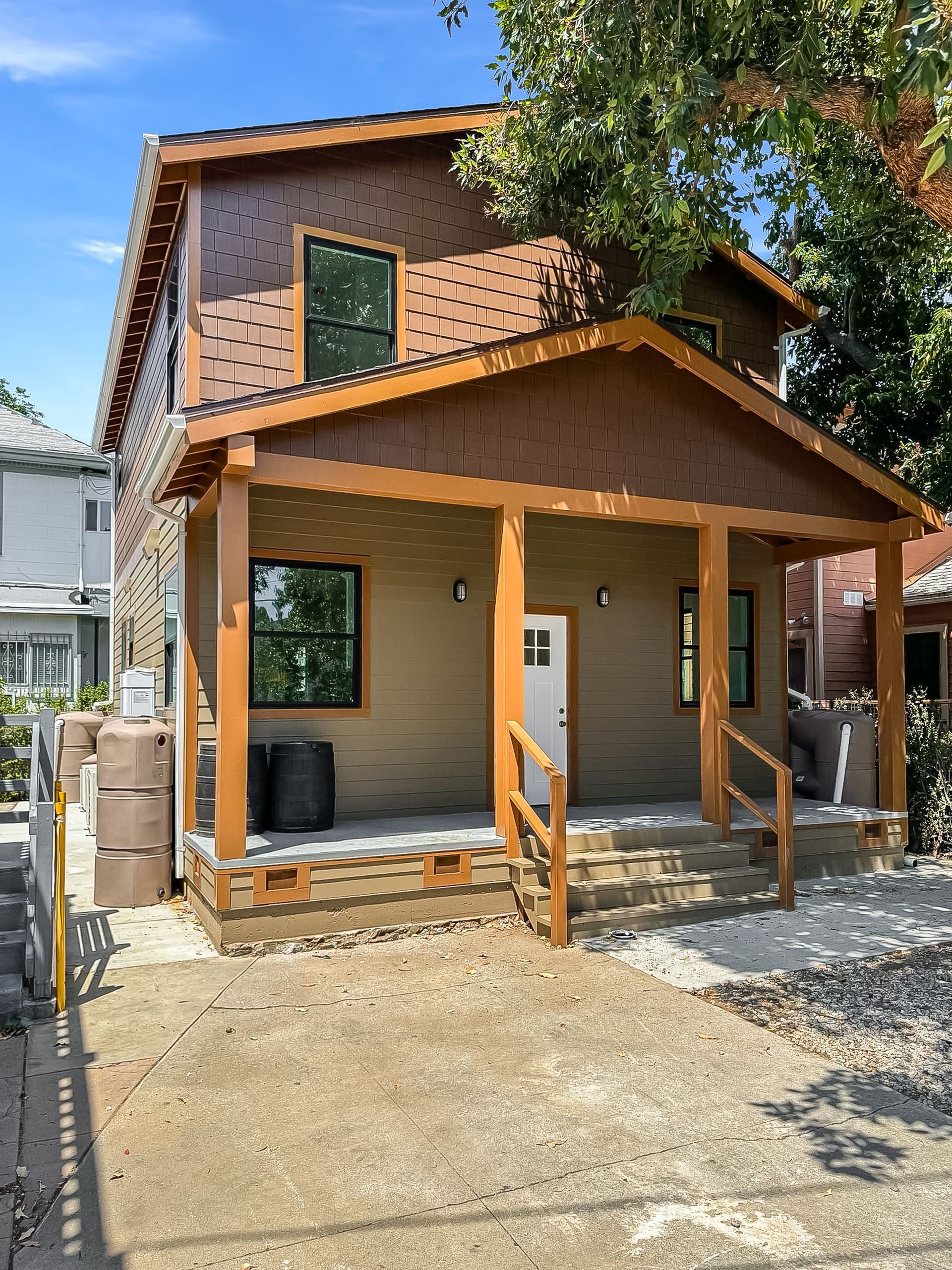 Two-story brown house with a covered porch and wooden railing, viewed from the front in a gravel driveway.