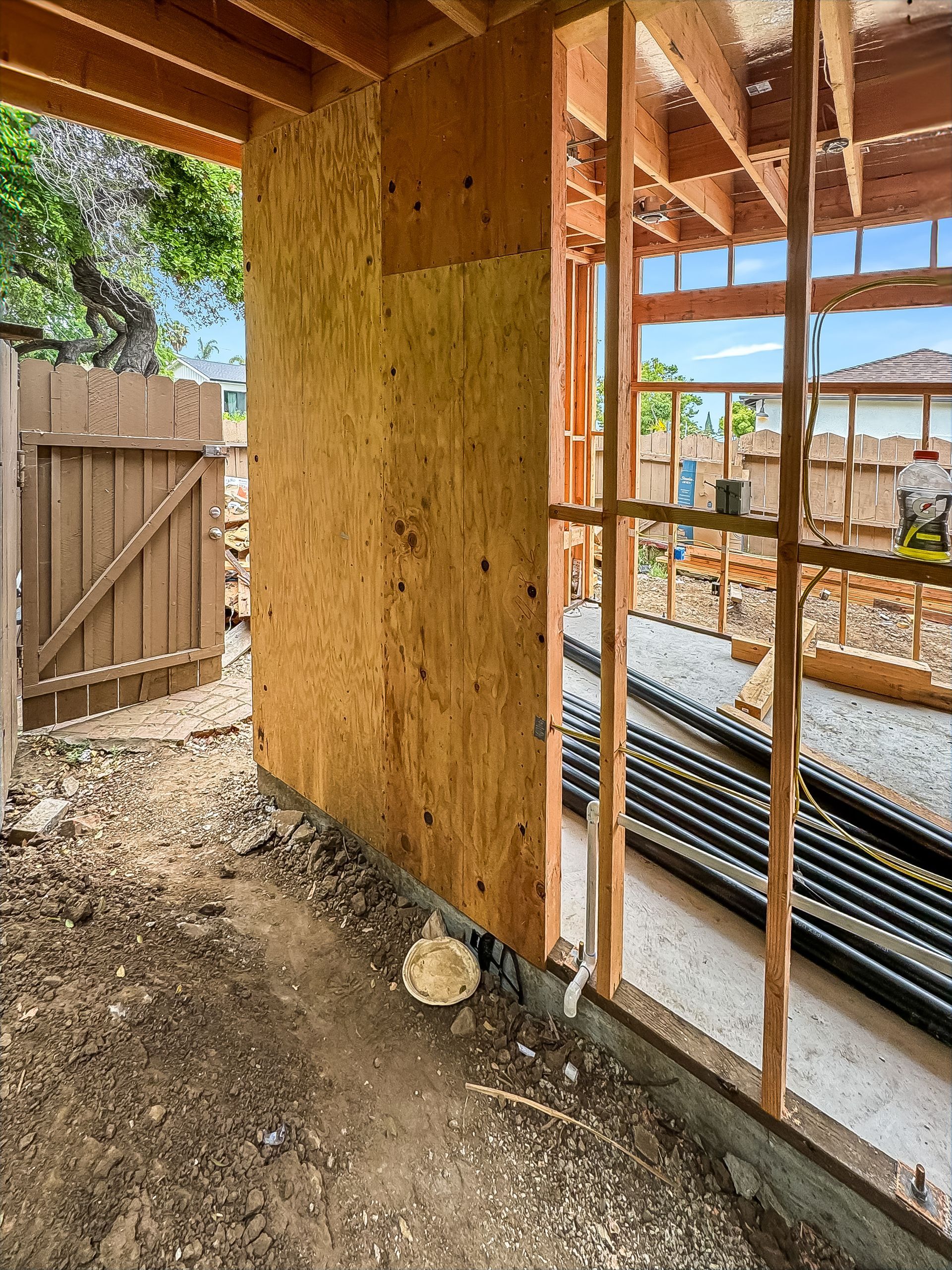 Partially framed house interior with plywood wall, exposed studs, and a dirt floor near a wooden gate