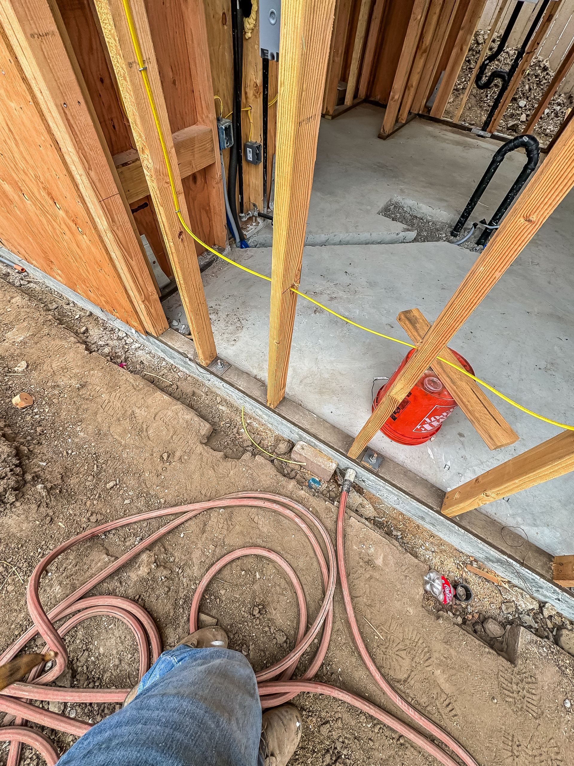 Construction site with exposed wall framing, concrete floor, coiled hoses, and a yellow caution line.