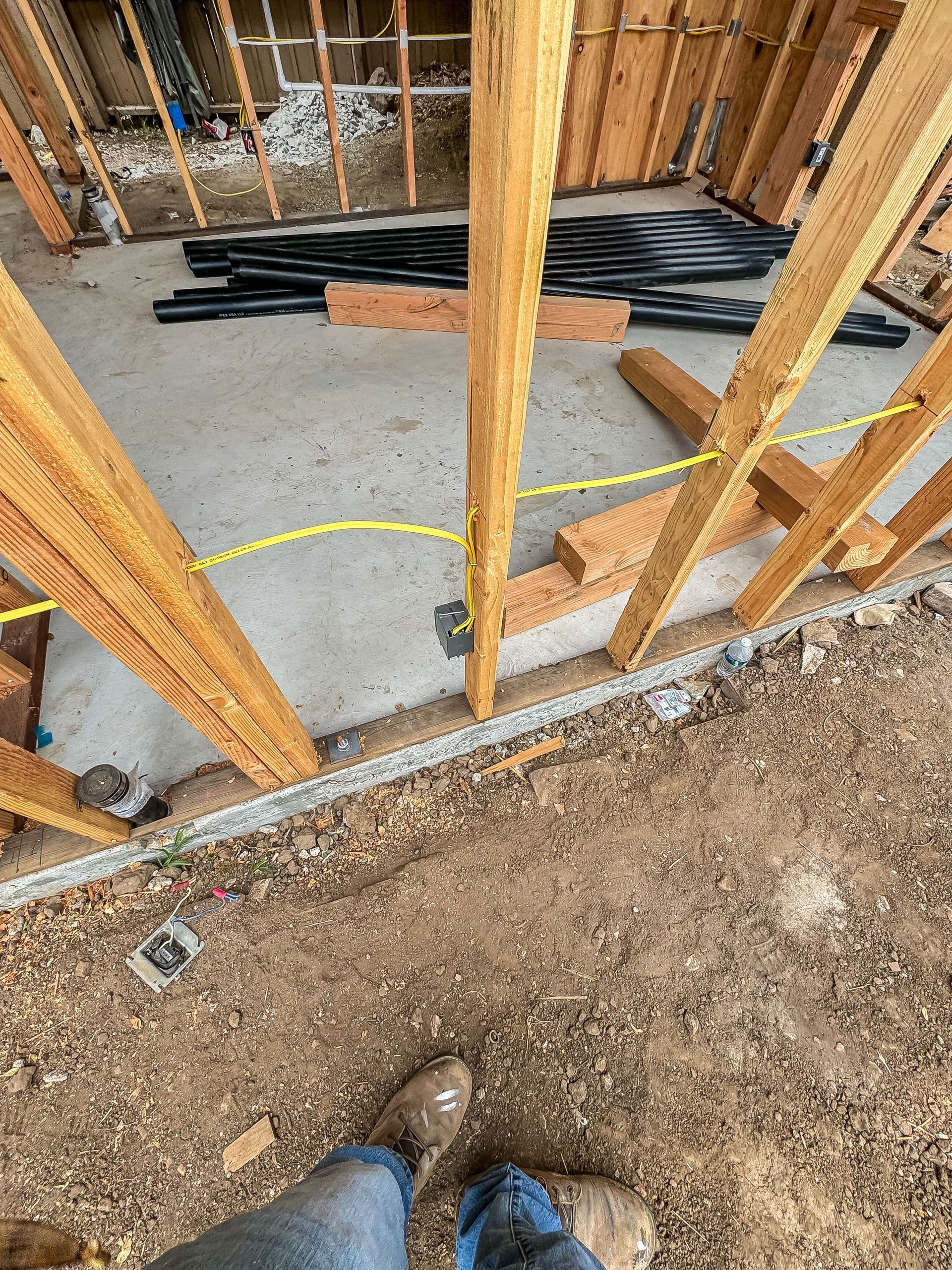 Framed construction site with concrete floor, stacked lumber, yellow hose, and a worker’s boots at bottom edge