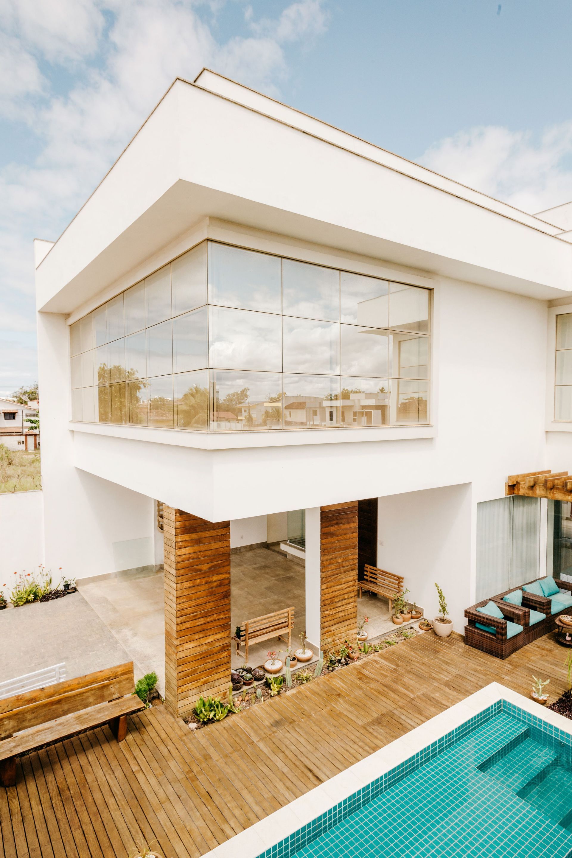 Modern white house with large glass windows, wooden deck, brick columns, and a turquoise pool in the foreground