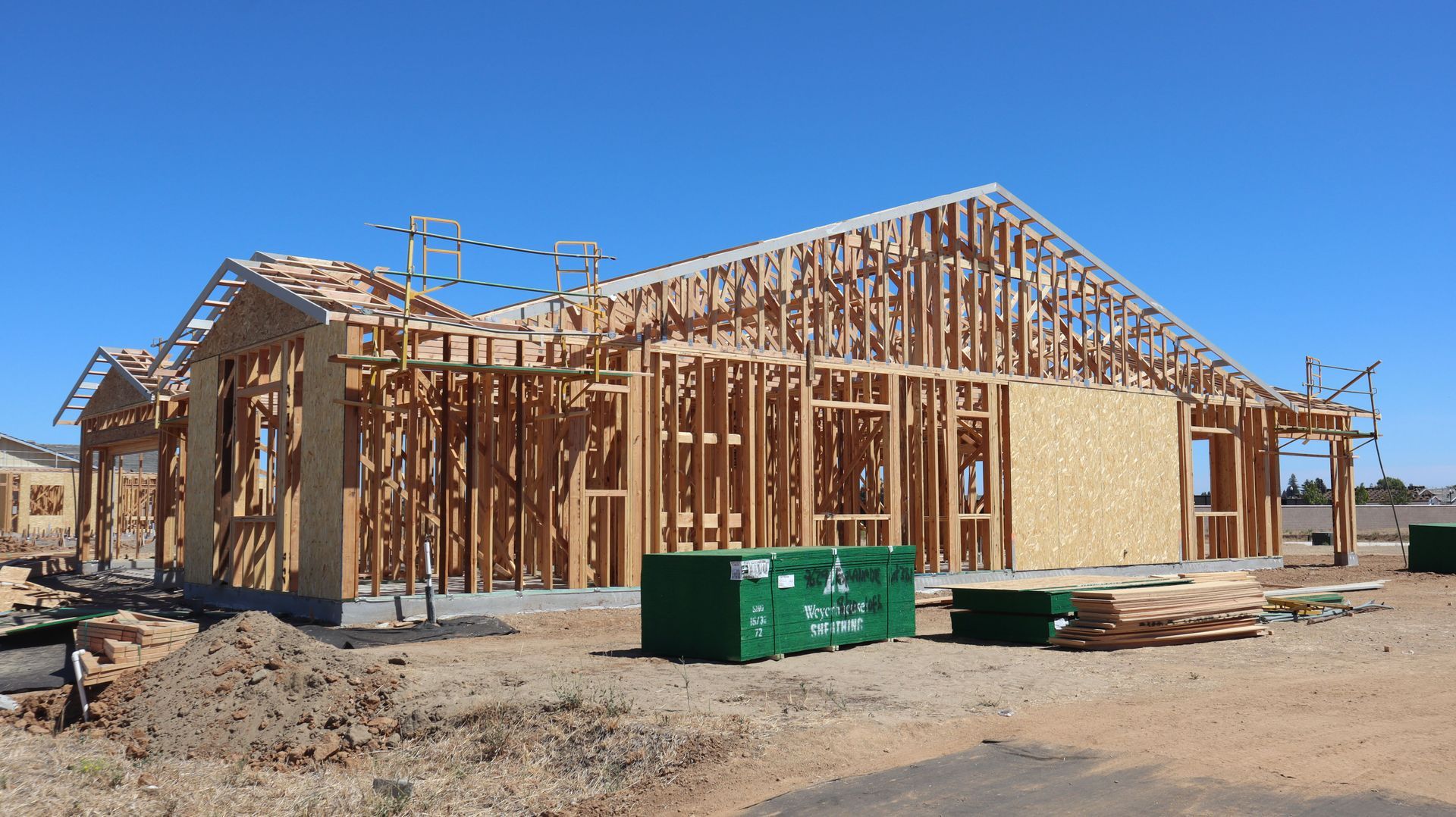 Wood-framed house under construction on a dirt lot, with green dumpsters in front and a blue sky.