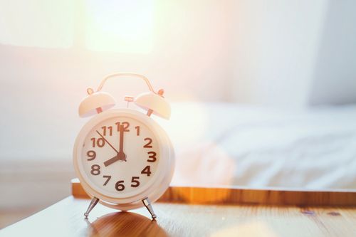 A white alarm clock is sitting on a wooden table next to a bed.