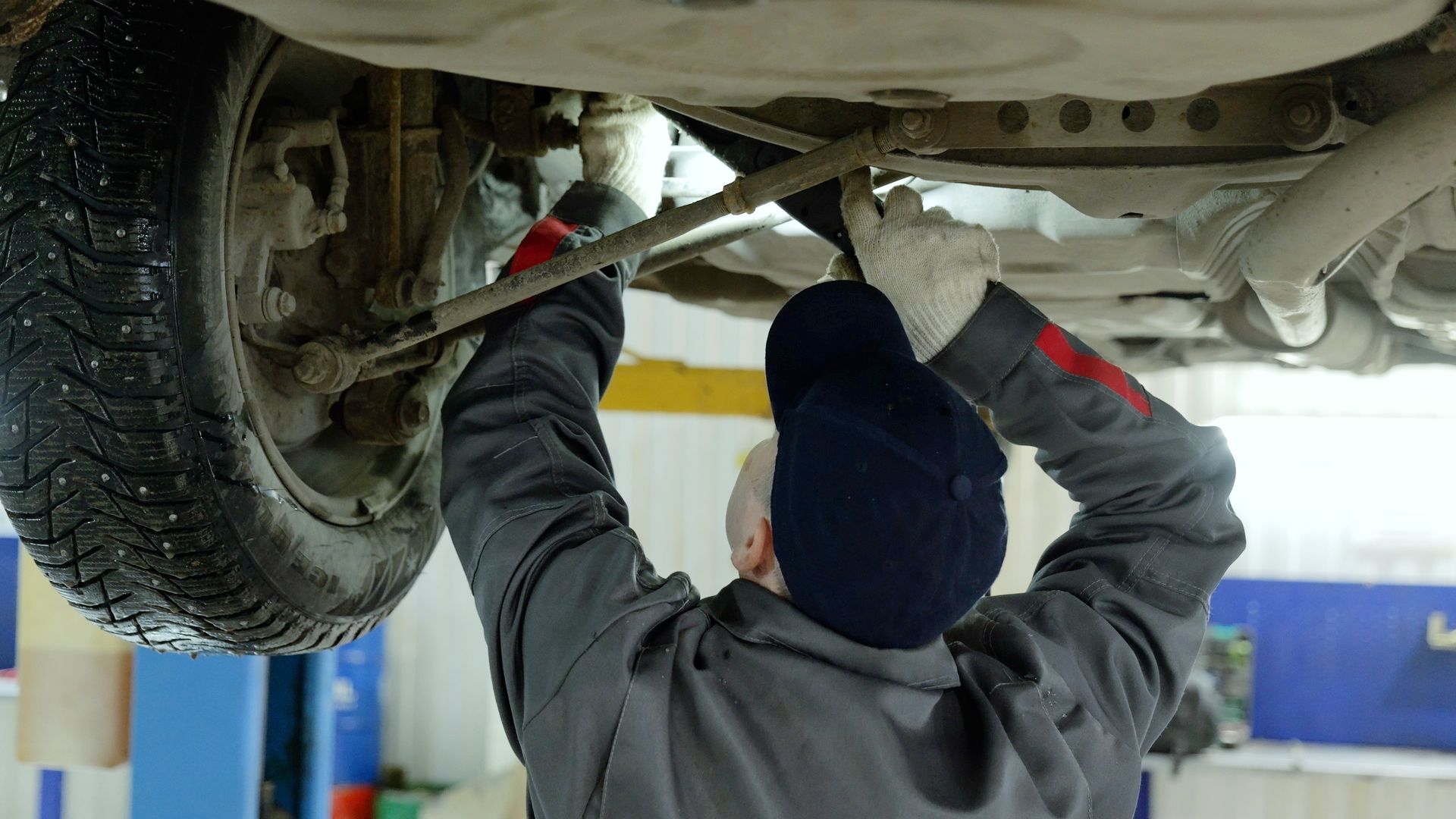 Mechanic inspecting the underside of a raised car in a garage, reaching up with both hands.
