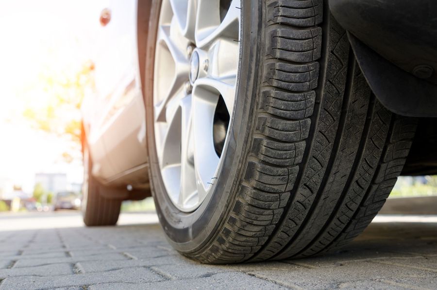 Close-up of a car tire and wheel on a sunlit road near a white vehicle.