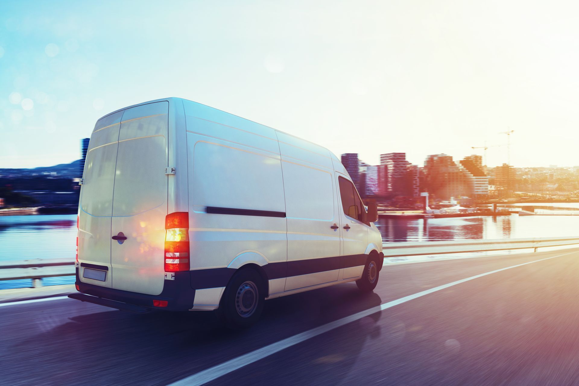 White delivery van driving on a wet road at sunset, with city lights reflected ahead