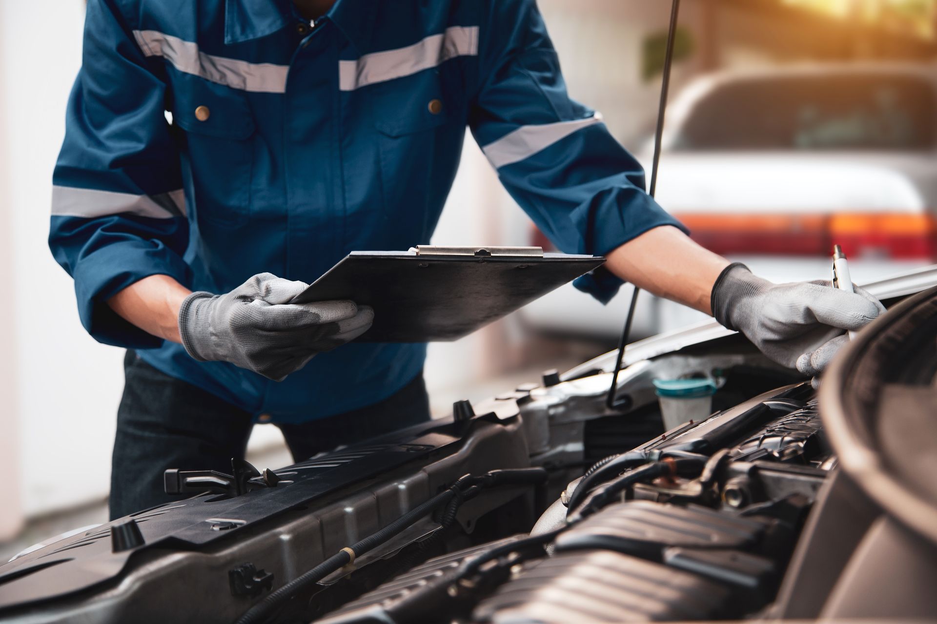 Mechanic in blue uniform inspecting a car engine with a clipboard in a garage