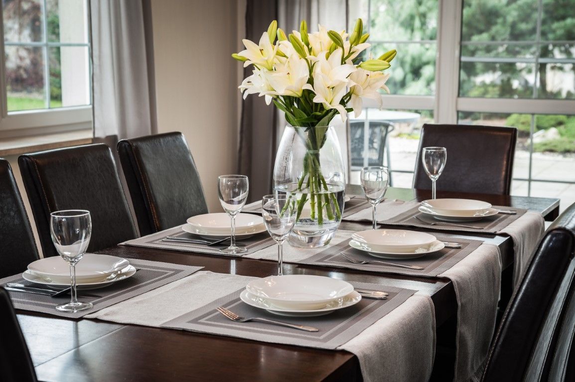 Set dining table with white plates, wine glasses, and a vase of white lilies by a window