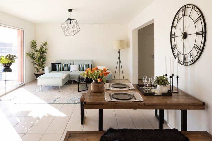 Bright living-dining room with a wooden table, white sofa, large wall clock, and flowers in a vase