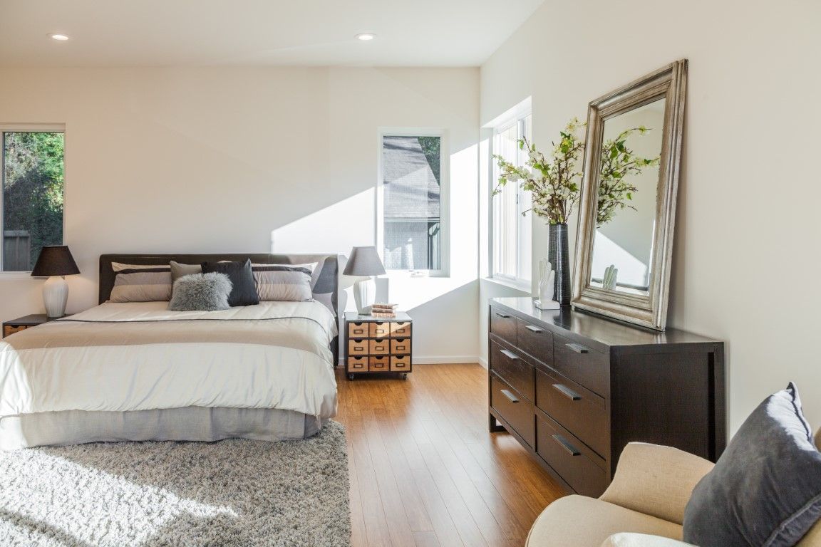 Bright bedroom with white bed, dark dresser, large mirror, and sunlight from windows