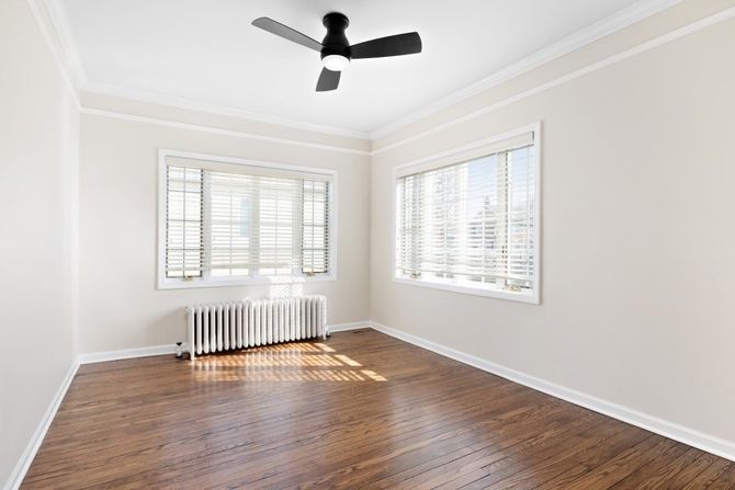Empty bright room with hardwood floors, white walls, windows, radiator, and a ceiling fan