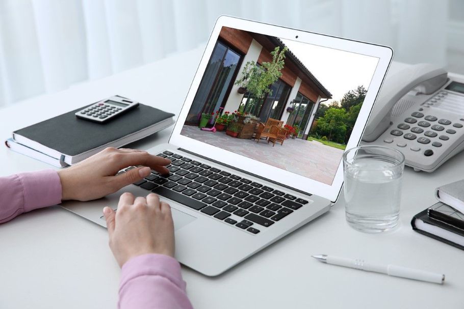 Hands typing on a laptop showing a house, with a notebook, phone, and glass on a clean desk