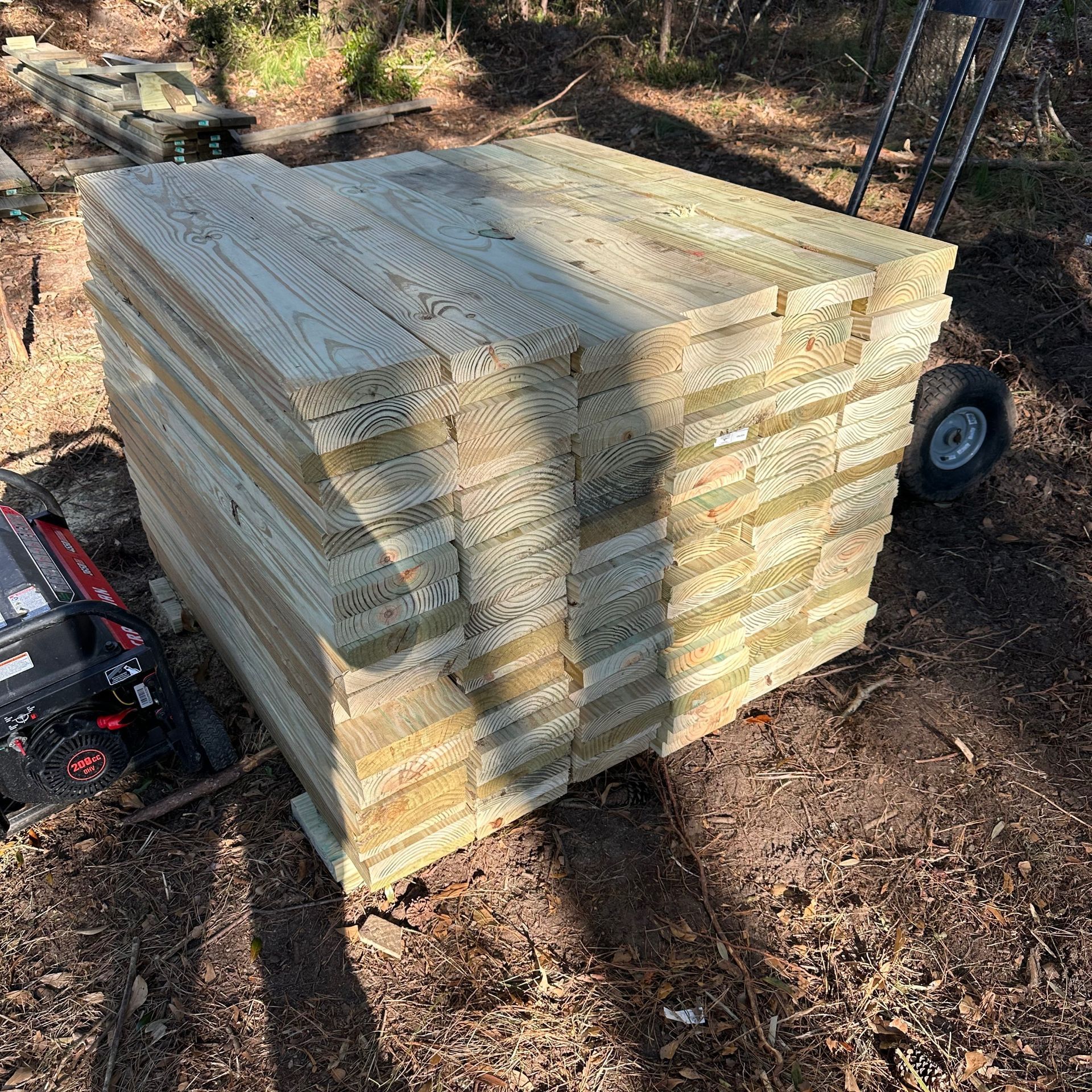 Stacked wooden boards on a pallet outdoors in a wooded area, with a cart beside them.