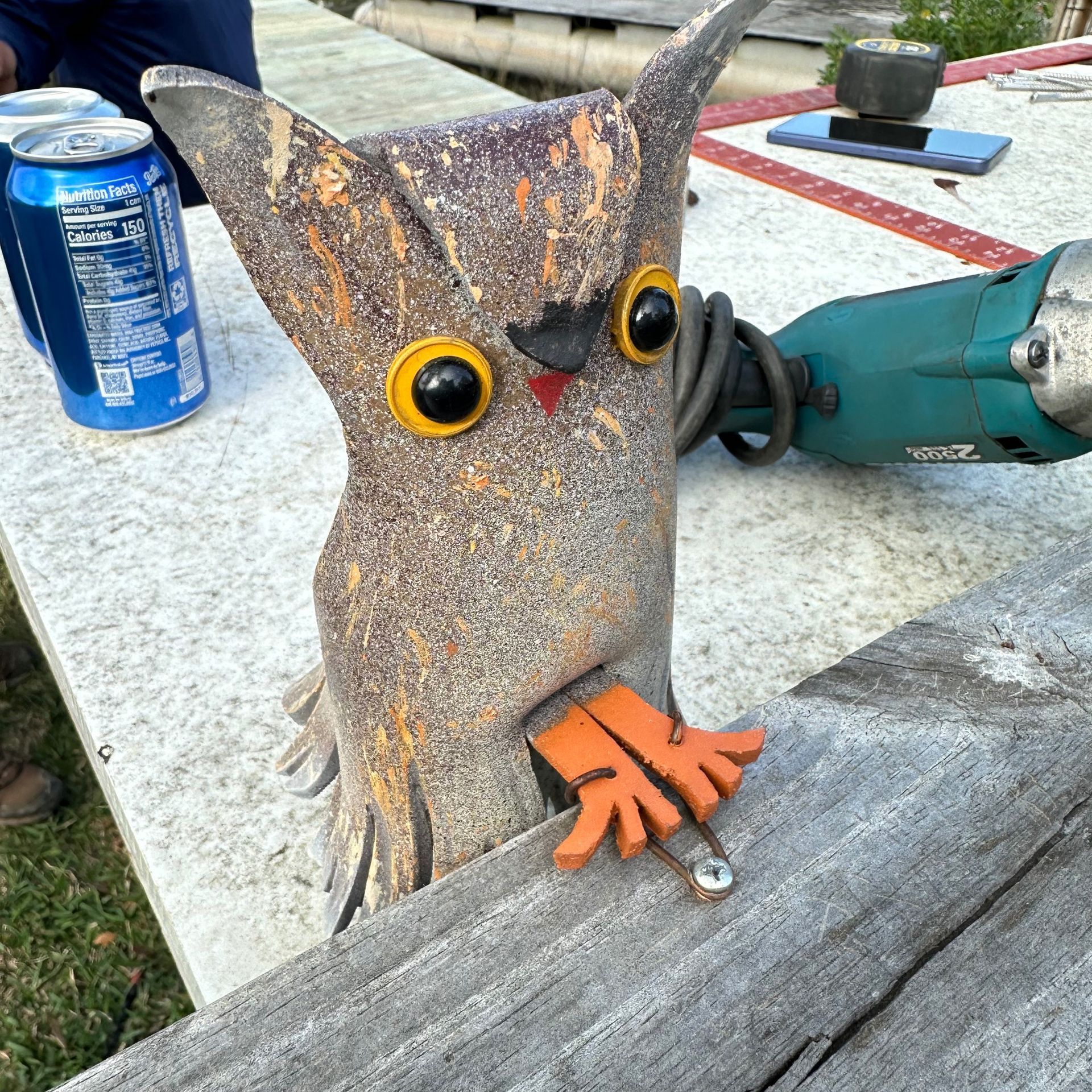 Rusty owl sculpture with orange feet on a tabletop, next to a blue soda can and a power tool.